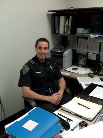 Sam wearing his police uniform sitting at his desk smiling at the camera